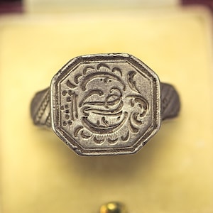 May include: A silver signet ring with an octagonal face featuring an engraved design of a stylized letter 'E' surrounded by floral motifs.