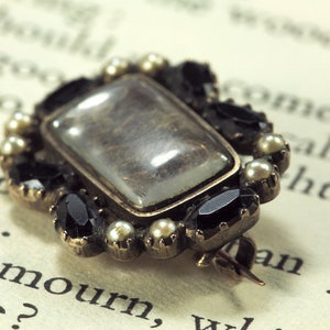 May include: A gold-toned brooch with a rectangular glass center, surrounded by black and white stones. The brooch is on a page of a book.