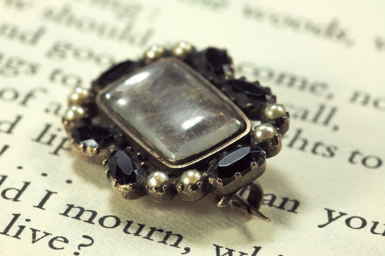 May include: A gold-toned brooch with a rectangular glass center, surrounded by black and white stones. The brooch is on a page of a book.