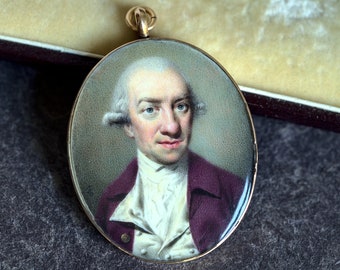 May include: A gold oval locket with a painted portrait of a man with white hair and a burgundy jacket. The man is wearing a white shirt with a ruffled collar.