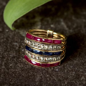 May include: A gold ring with multiple bands, each featuring a different design. Some bands have red gemstones, others have clear stones, and one has blue gemstones. The ring is set against a dark, textured background.
