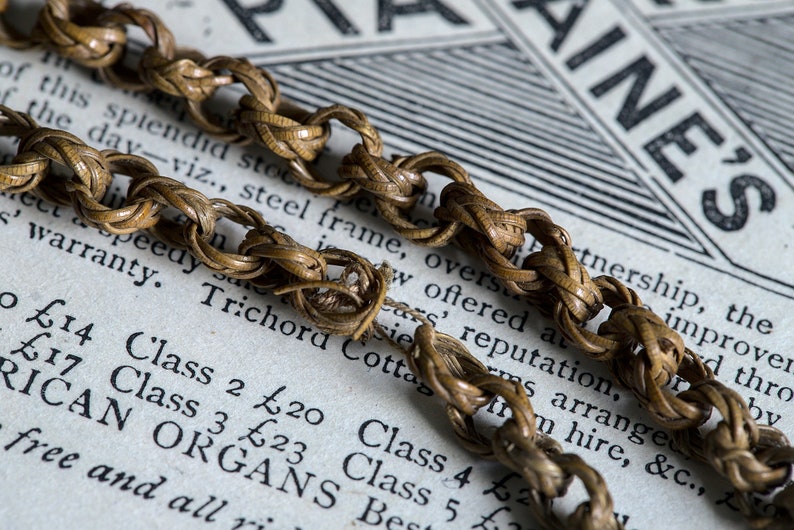 May include: A close-up of a woven chain made of brown material. The chain is lying on top of a piece of paper with text that reads "Class 2 &pound;20 Class 4 &pound;23 Class 5 &pound;25 Best" and "RICAN ORGANS".
