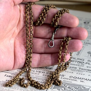 May include: A gold chain with a silver clasp. The chain is lying on a newspaper with text that includes the words "Sterling Silver", "Electro-Plate", "Highest Quality", "Trade Prices", "London, W.", and "Belfast".