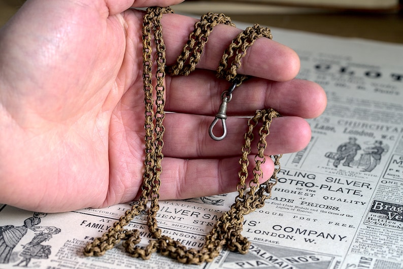 May include: A gold chain with a silver clasp. The chain is lying on a newspaper with text that includes the words "Sterling Silver", "Electro-Plate", "Highest Quality", "Trade Prices", "London, W.", and "Belfast".
