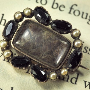 May include: A silver locket brooch with a glass insert containing a lock of hair. The locket is surrounded by a ring of black and white gemstones.