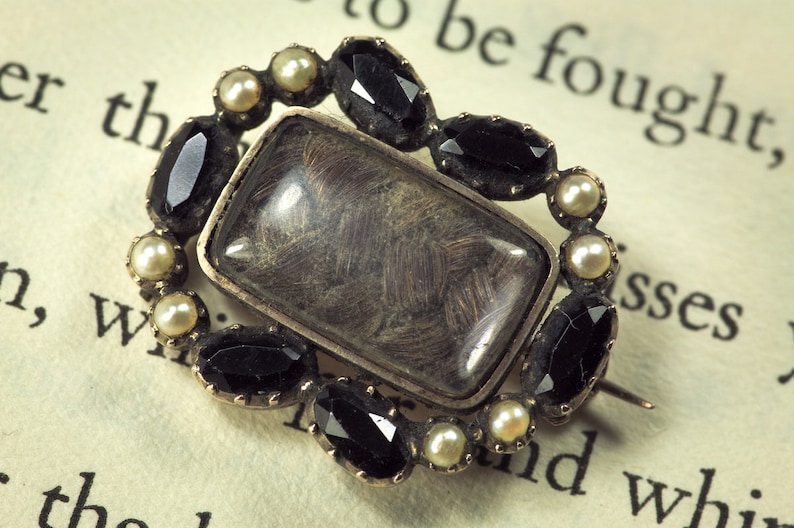 May include: A silver locket brooch with a glass insert containing a lock of hair. The locket is surrounded by a ring of black and white gemstones.
