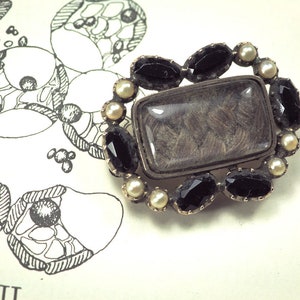 May include: A silver brooch with a rectangular glass locket containing a lock of hair. The locket is surrounded by a border of black gemstones and white pearls.