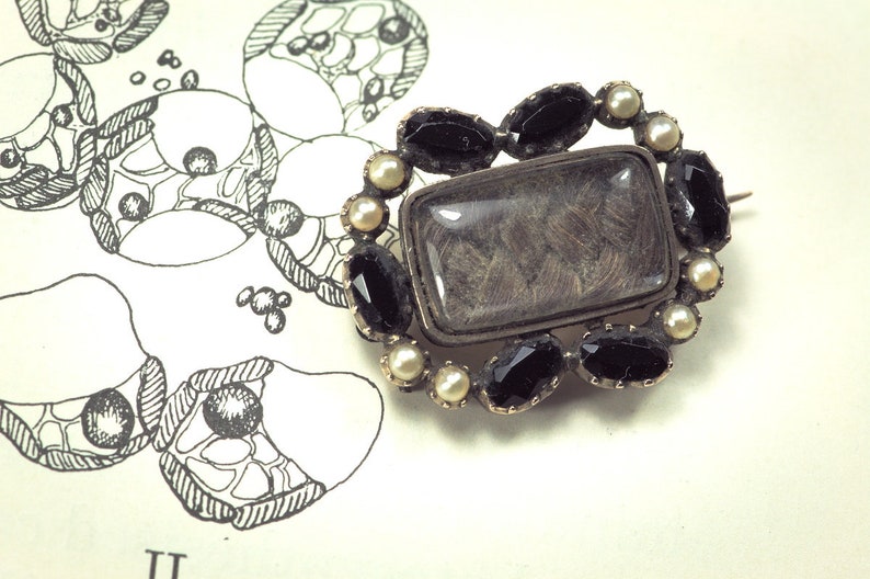 May include: A silver brooch with a rectangular glass locket containing a lock of hair. The locket is surrounded by a border of black gemstones and white pearls.