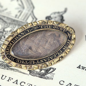 May include: A gold-toned oval locket with a black enamel border and inscription "REMEMBRANCE OF THE JUST",  "THE PLEASANTIS THE MOST", and "FLOW" around the edge. The locket contains a woven lock of hair inside a glass oval.