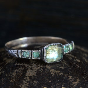 Puede incluir: Un anillo de plata con una piedra preciosa grande, facetada, de color verde claro, rodeada de cuatro piedras preciosas verdes más pequeñas.