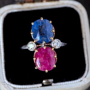 May include: A ring with a large blue sapphire and a smaller pink ruby, both set in gold. The ring also has two smaller diamonds on either side of the ruby.
