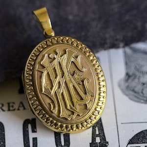 May include: A gold locket with a detailed engraved monogram. The locket has a decorative border and a loop for a chain.