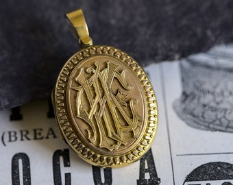 Könnte beinhalten: Ein goldenes Medaillon mit einem detailliert gravierten Monogramm. Das Medaillon hat einen dekorativen Rand und eine Schlaufe für eine Kette.