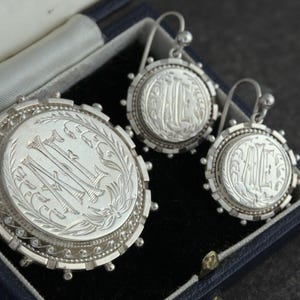 May include: A set of silver earrings and a brooch with engraved initials. The jewelry is displayed in a blue velvet box.