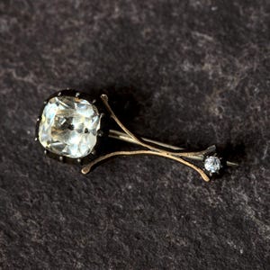 May include: An antique brooch featuring a large, square-cut, clear gemstone set in a dark metal frame. The brooch has a delicate, branch-like design with a smaller, round gemstone at the end of one branch. The setting is gold-toned.