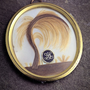May include: A gold-framed locket with a hair art design of a weeping willow tree. The tree is made of brown hair and the background is white. The locket has a black circle with the initials "H.L." in gold lettering.