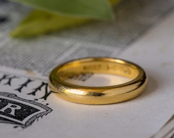 May include: A gold wedding band with a smooth, rounded design. The ring is set against a backdrop of aged paper and a blurred green leaf. The ring's surface reflects light, highlighting its polished finish.