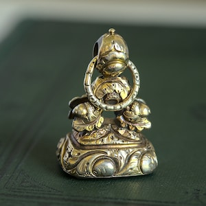 May include: A gold-toned metal seal with intricate floral designs and a ring at the top. The seal is on a green surface.