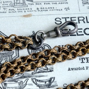 May include: A close-up of a braided chain with a silver clasp. The chain is made of woven metal and has a classic design. The clasp is a simple hook and eye style.