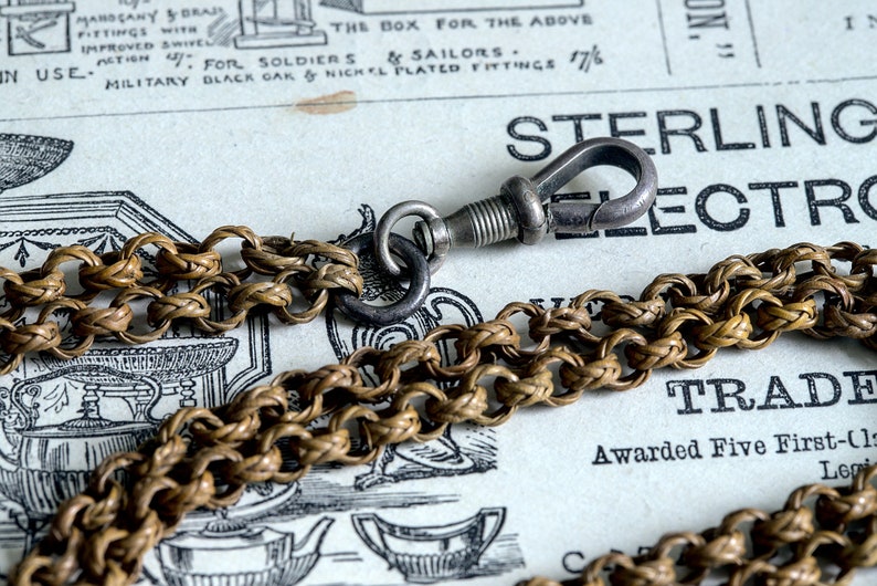 May include: A close-up of a braided chain with a silver clasp. The chain is made of woven metal and has a classic design. The clasp is a simple hook and eye style.