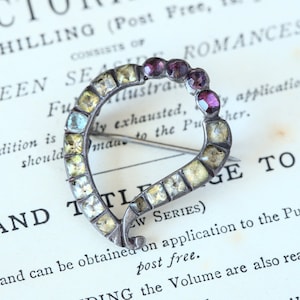 May include: A heart-shaped brooch with a silver setting and a variety of colorful stones. The stones are set in a square shape and are arranged in a pattern. The brooch has a pin on the back for attaching it to clothing.