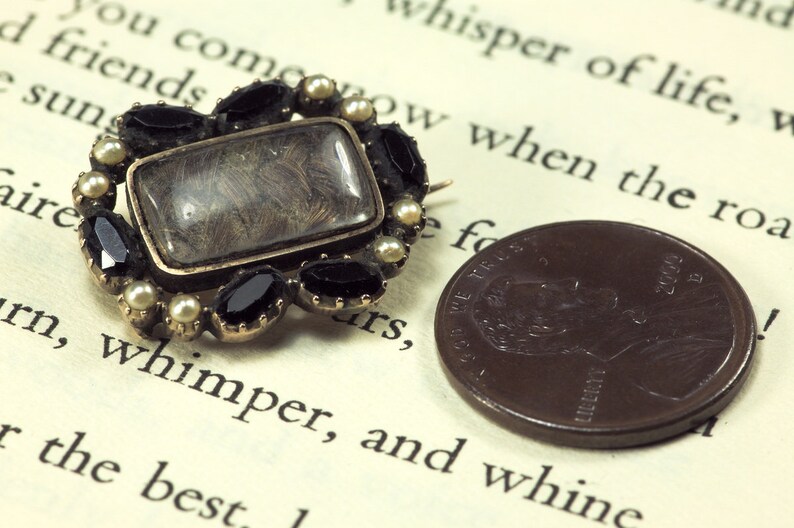 May include: A gold-toned locket with a glass window containing a lock of hair. The locket is surrounded by black and white gemstones.