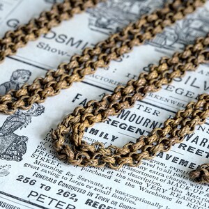 May include: A close-up of a gold chain necklace with a rope-like design. The chain is lying on a vintage newspaper with text that includes the words "Family Mourning Warehouse".