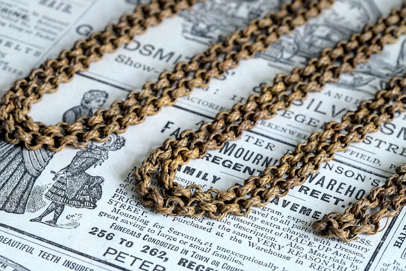 May include: A close-up of a gold chain necklace with a rope-like design. The chain is lying on a vintage newspaper with text that includes the words "Family Mourning Warehouse".