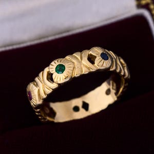 Op de afbeelding: Een gouden ring met een herhalend bloemmotief, elke bloem geaccentueerd met een kleine edelsteen. De edelstenen zijn rood, groen en blauw. De ring wordt gepresenteerd in een bordeauxrode sieradendoos.