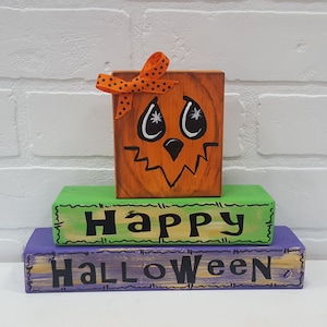 Könnte beinhalten: Ein Halloween-Holzblock-Set. Der obere Block ist orange und als Kürbislaterne mit einer Schleife mit Punkten geschnitzt. Der mittlere Block ist grün mit dem Wort "HAPPY" in Schwarz. Der untere Block ist lila mit "HALLOWEEN" in Schwarz.