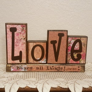 May include: Wooden block sign spelling "LOVE" with floral and brown accents. The sign includes the text "Bears all things! 1Cor 13:7". The letters are in a dark brown font.