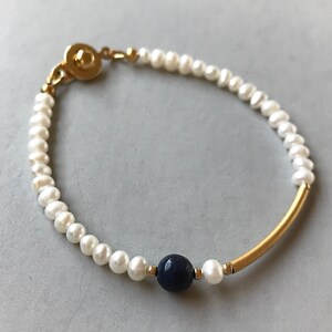 May include: A bracelet featuring a strand of small white pearls, a single round dark blue faceted bead, a curved gold-tone metal tube, and small gold-tone spacer beads. The piece is secured with a round gold-tone metal clasp closure.