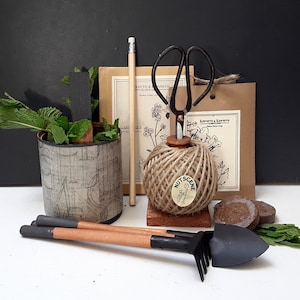 May include: A gardening kit containing a small pot of green plants, a ball of twine, a pair of scissors, a small trowel, a pencil, and two seed starter discs. The twine ball has a label that says "Nut Scene".