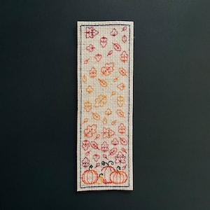 May include: A rectangular bookmark with a beige background and a black border. The design features embroidered autumn leaves in shades of orange and red, with pumpkins at the bottom. The bookmark is approximately 2 inches wide and 8 inches long.