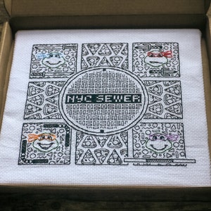 Blackwork embroidery of four turtles, each with a different colour eye mask. Pattern also shows a central manhole cover design reading 'NYC Sewer', and four panels have a recurring pizza motif.