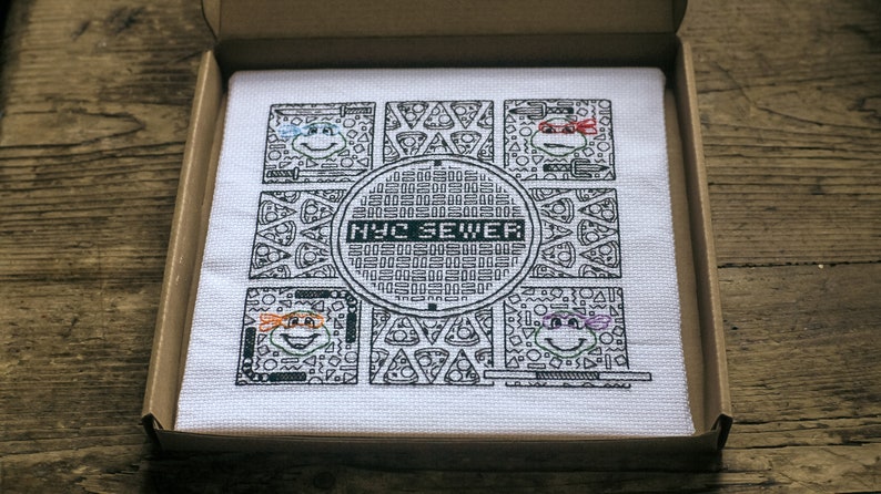 Blackwork embroidery of four turtles, each with a different colour eye mask. Pattern also shows a central manhole cover design reading 'NYC Sewer', and four panels have a recurring pizza motif.