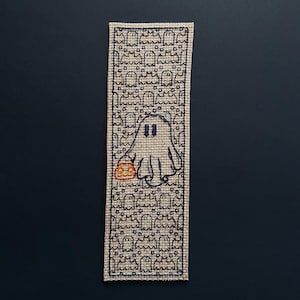 May include: A beige fabric bookmark featuring a black outline of a ghost holding a small orange pumpkin. The background is filled with a pattern of smaller ghosts.