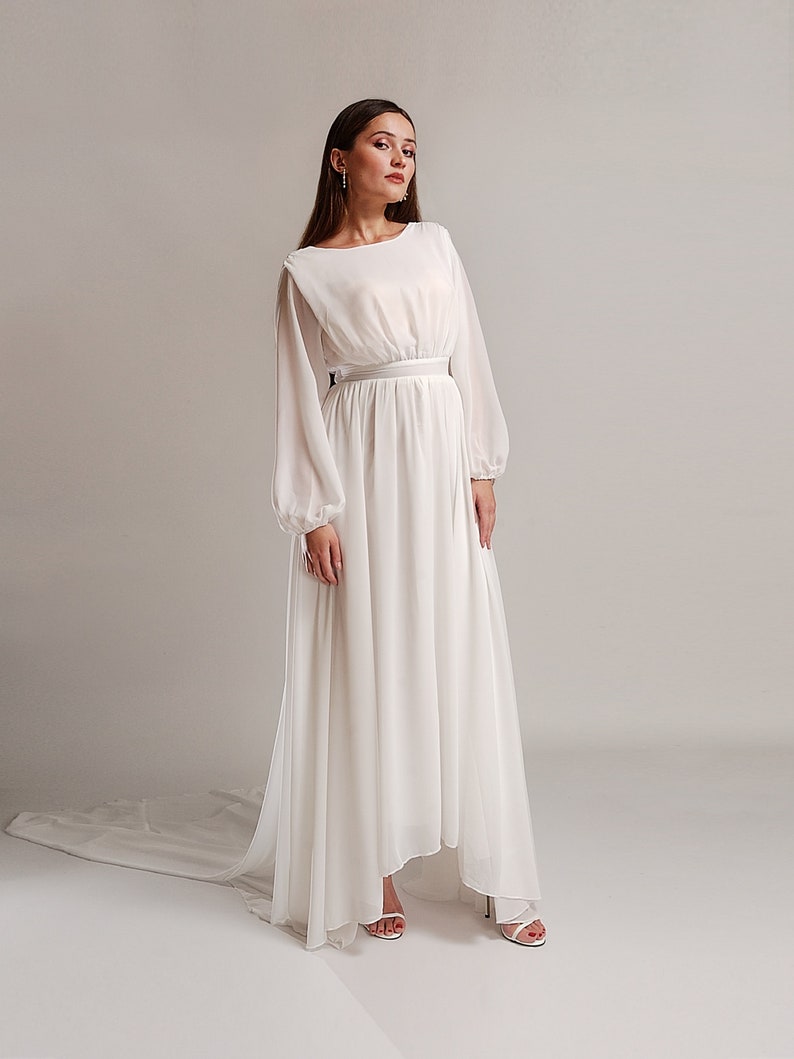 May include: A white maxi dress with long sleeves and a fitted waist. The dress has a flowing skirt and a high neckline.