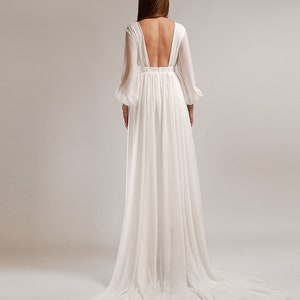 May include: A white, flowing wedding dress with a deep V-neck back and long sleeves. The dress has a flowing skirt and a train.