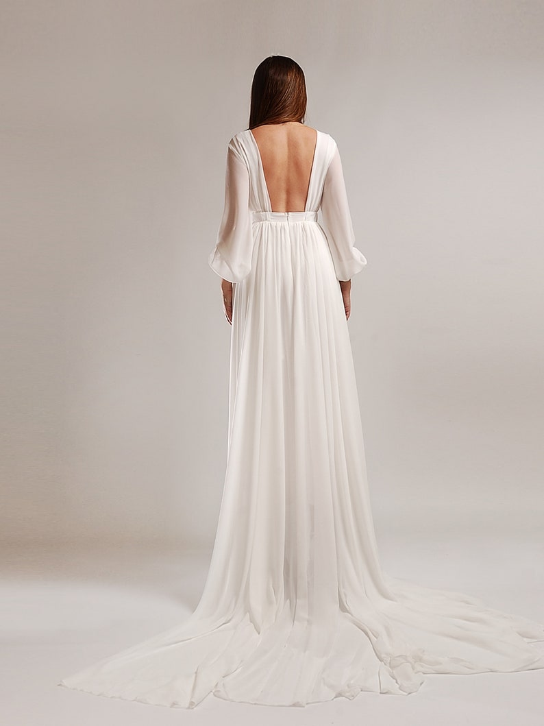 May include: A white, flowing wedding dress with a deep V-neck back and long sleeves. The dress has a flowing skirt and a train.