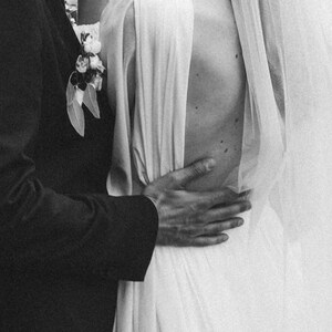 May include: A black and white photo of a couple kissing. The woman is wearing a white wedding dress with a veil and the man is wearing a black suit. The couple is standing in front of a green leafy background.