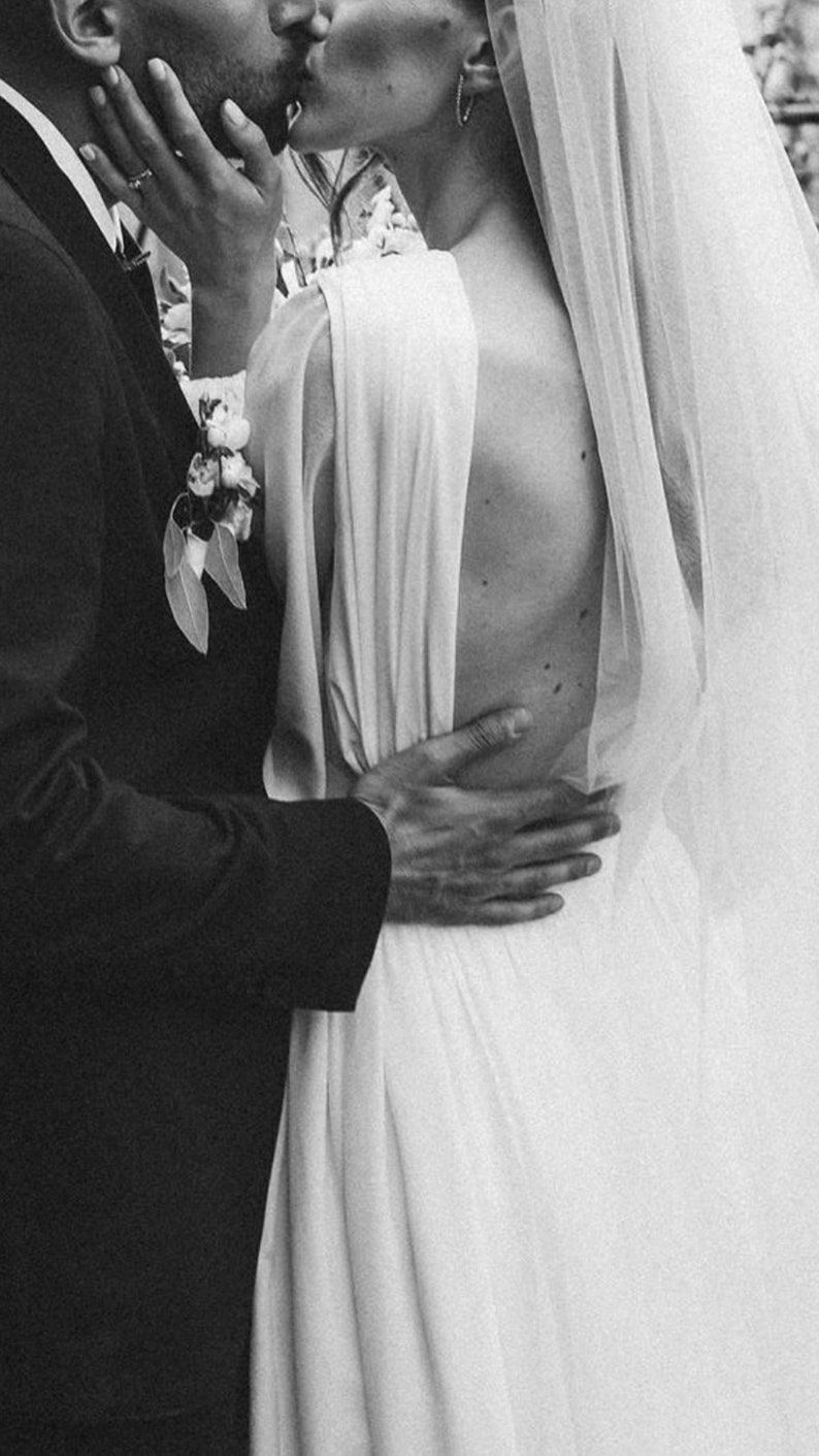 May include: A black and white photo of a couple kissing. The woman is wearing a white wedding dress with a veil and the man is wearing a black suit. The couple is standing in front of a green leafy background.