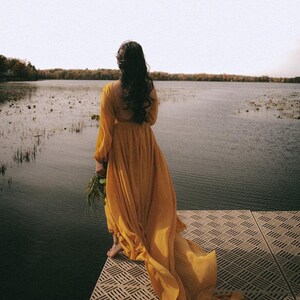 May include: A woman in a long, flowing yellow dress stands on a wooden dock overlooking a lake. The dress has a long train that trails behind her. The lake is calm and still, and the sky is a soft blue.