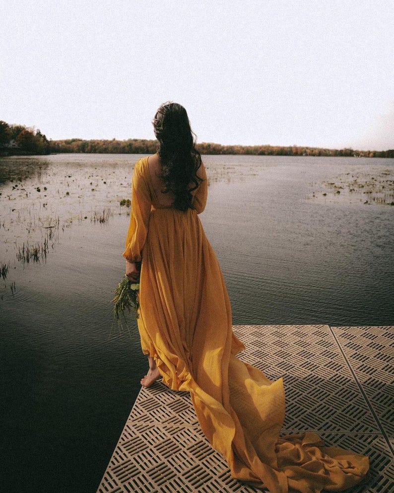 May include: A woman in a long, flowing yellow dress stands on a wooden dock overlooking a lake. The dress has a long train that trails behind her. The lake is calm and still, and the sky is a soft blue.