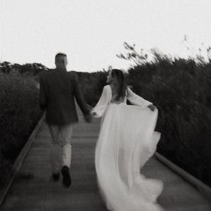 May include: A black and white photo of a couple walking away from the camera on a path. The woman is wearing a white dress and the man is wearing a suit jacket and pants.