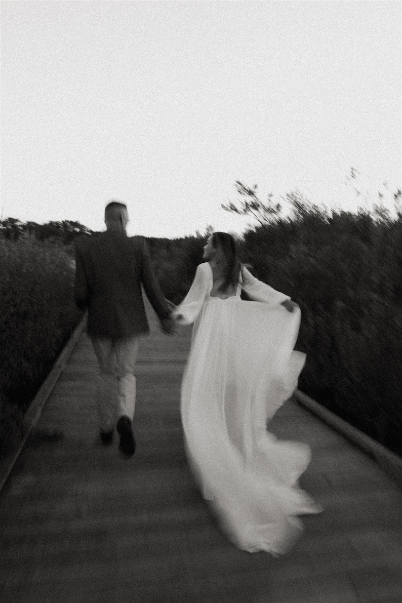 May include: A black and white photo of a couple walking away from the camera on a path. The woman is wearing a white dress and the man is wearing a suit jacket and pants.