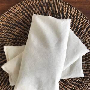 May include: Two white fabric eye pillows on a woven brown wicker surface.