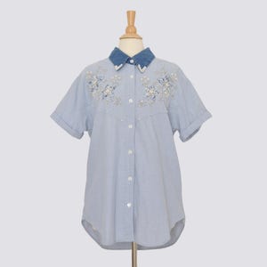 May include: Light blue short-sleeved shirt with a denim collar. The shirt features floral embroidery with sequins and pearl accents around the neckline. The shirt is button-down and has a curved hemline.
