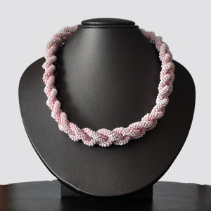 May include: A pink and white beaded necklace with a twisted design. The necklace is displayed on a black mannequin bust.