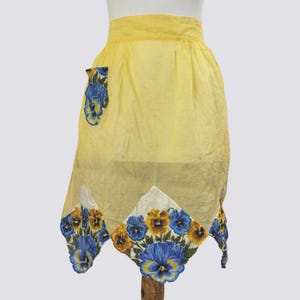 May include: A yellow apron with a blue and yellow floral print. The apron has a single pocket and a scalloped hem.